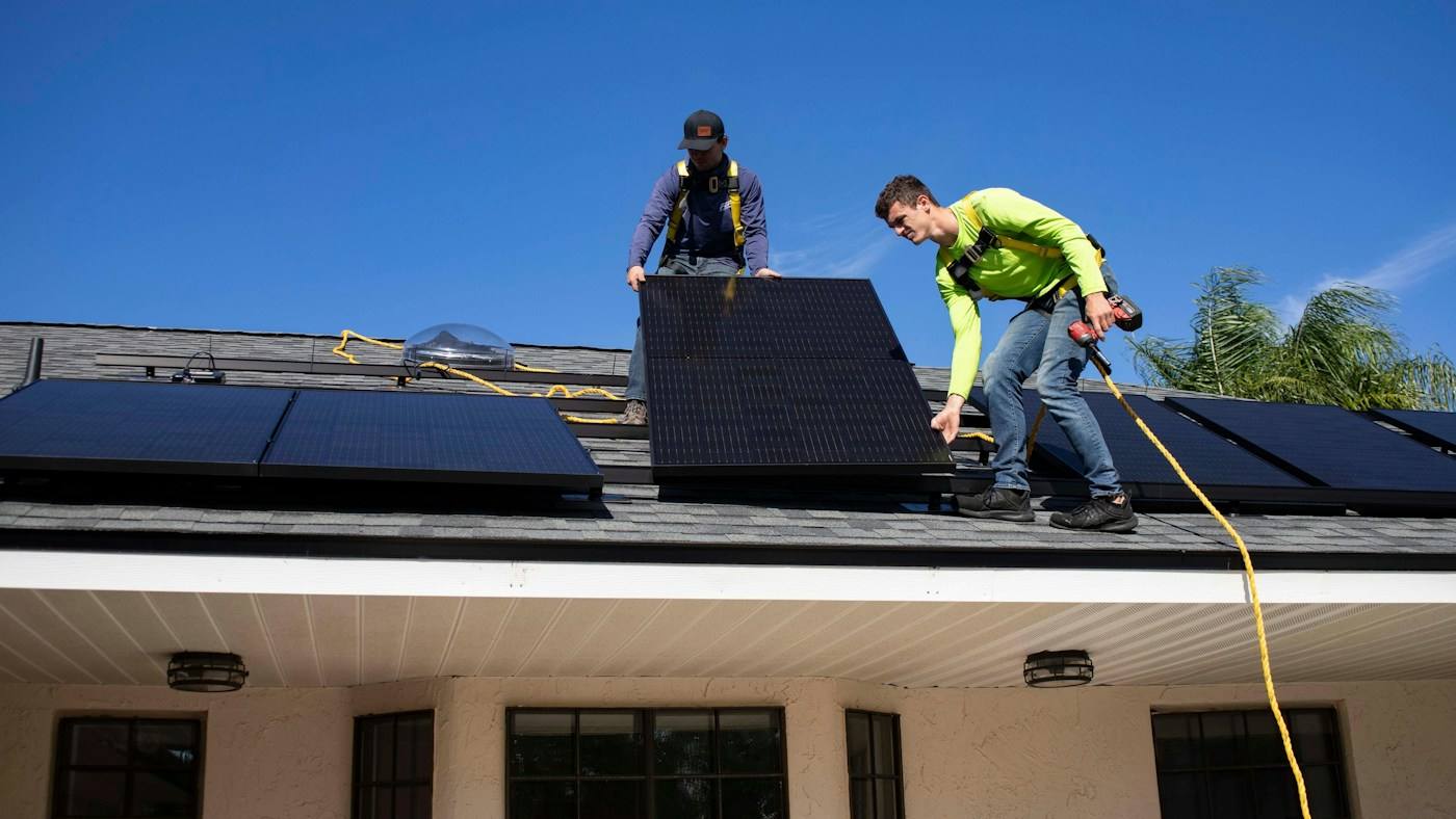 Dois instaladores com cinto de segurança fixando painéis solares em um telhado.