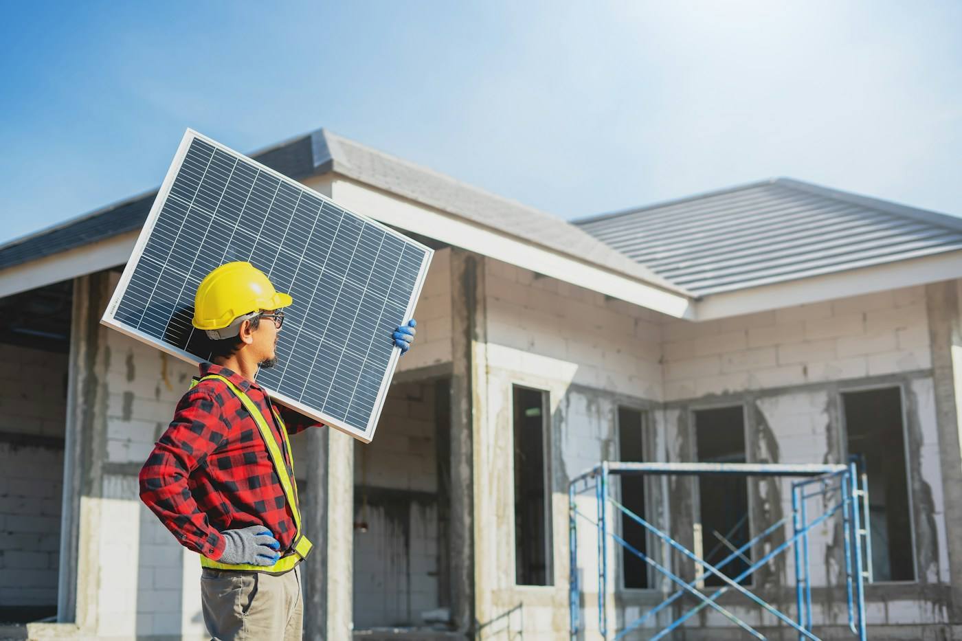 Instalador de capacete carregando um painel solar em frente a uma construção.